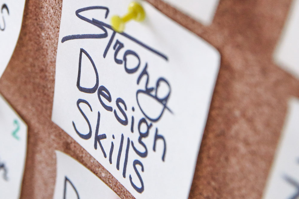 Photo of sticky note that says Strong Design Skills pinned on corkboard.